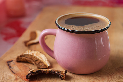 Mushroom coffee chaga superfood. Dried mushrooms and fresh brewed pink cup of hot drink with foam on wooden background vertical photo.Healthy organic energizing adaptogen,endurance boosting food trend